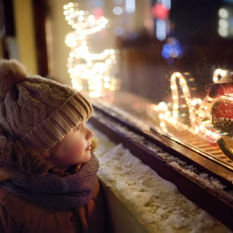 Little boy admires Christmas decorations in showcase of shop on winter evening. Tourist looking on Xmas toys and accessories on traditional Christmas market in Tallinn.