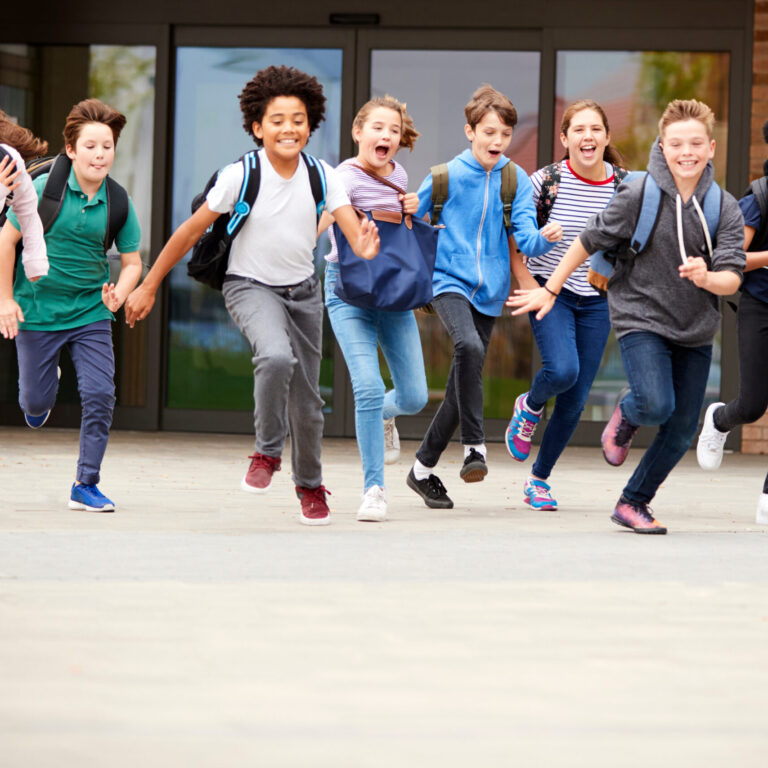 Group Of High School Students Running Out Of School Buildings Towards Camera At The End Of Class